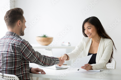 Happy asian businesswoman handshaking successful caucasian candidate holding employment agreement offering job contract, smiling friendly employer shaking new hire hand, diverse partners making deal