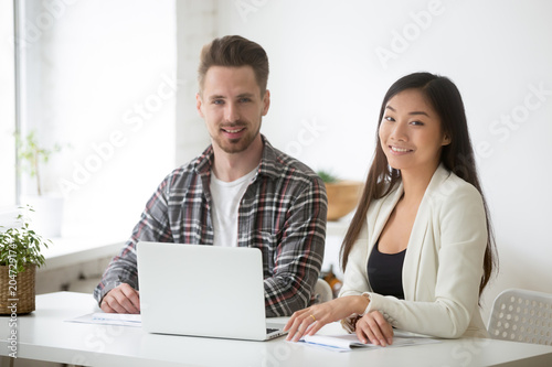 Smiling caucasian businessman and asian businesswoman looking at camera, diverse interns managers posing at workplace sitting in front of laptop, young motivated business professionals team portrait