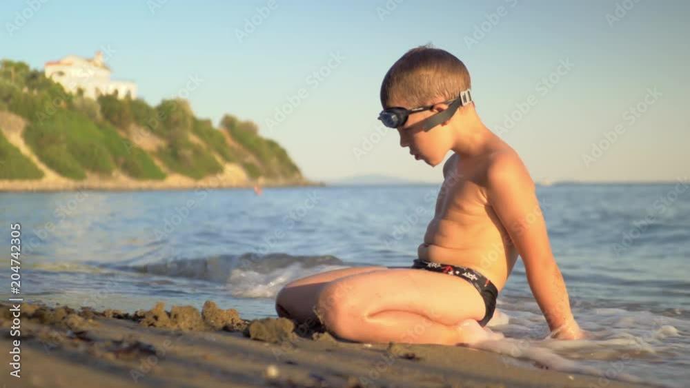 Boy sitting at the beach back to the sea and enjoying waves washing him, scene at sunset. Having great summer holidays at the seaside