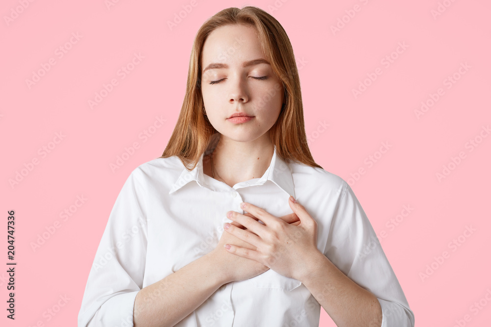 Pretty teenage girl keeps hands on chest and eyes closed, being thankful and grateful, expresses her devotion, wears shirt, isolated over pink background. My heart is for you! Friendly looking woman Pretty teenage girl keeps hands on chest and eyes closed, being thankful and grateful, expresses her devotion, wears shirt, isolated over pink background. My heart is for you! Friendly looking woman