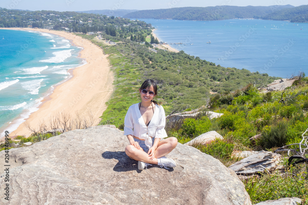 Young woman with sunglasses sits on the background of Palm beach on top of a rock at the lighthouse in Barrenjoey, Sydney, Australia.
