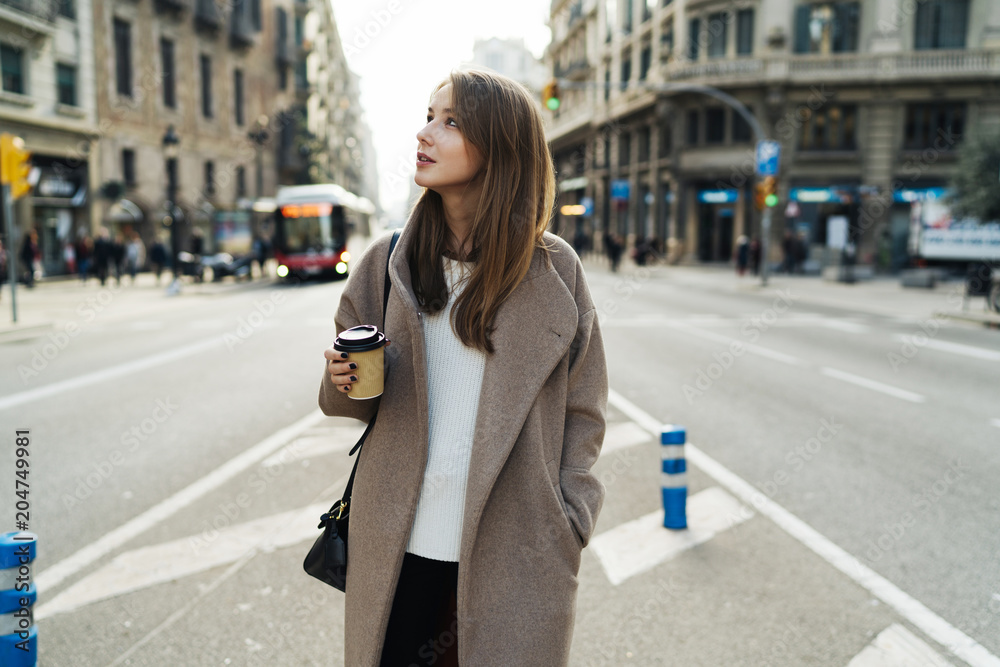Fototapeta premium Stylish tourist girl exlporing a city early in the morning with a paper coffee cup in her hand. Office employee enjoying after lunch walk time to the office with a take away coffee.