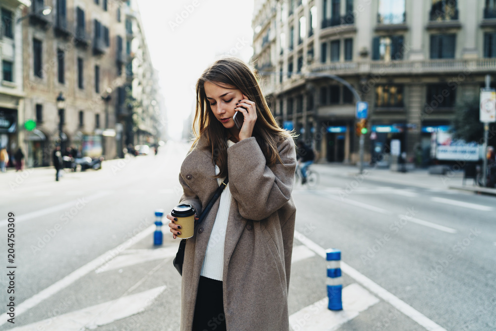 Fototapeta premium Successful business woman making business calls by a mobile phone to the partners while going to office with coffee. Fashionable hipster girl wearing stylish clothes chatting with friend by smartphone