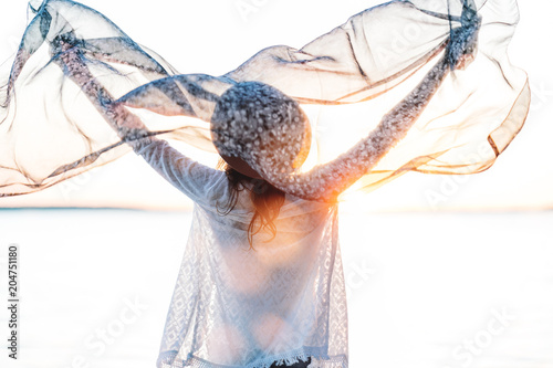 Beautiful boho girl standing with cloth in hand on summer breeze. Caucasian young female on beach with silk scarf looking at horizon