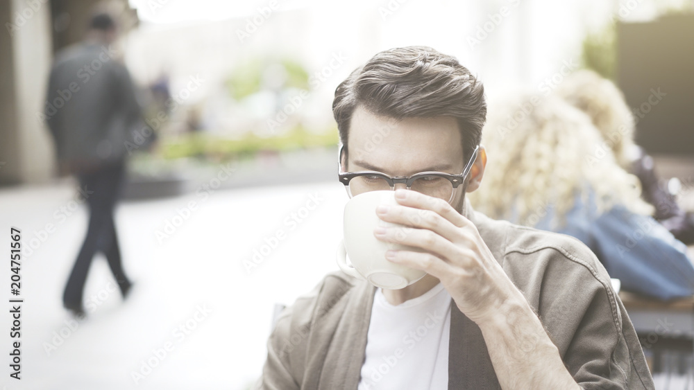 A closeup of handsome man wearing eyeglasses sitting at cafe drinking coffee outdoors A closeup of handsome man wearing eyeglasses sitting at cafe drinking coffee outdoors