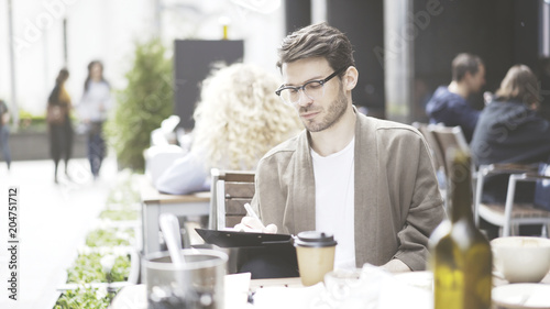 A handsome thoughtful pensive man in eyeglasses making notes at cafe outdoors