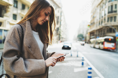 Successful freelancer typing email on  mobile phone connected to wifi while walking the street. Charming hipster girl wearing stylish clothes searching needed address in the internet on a smartphone.