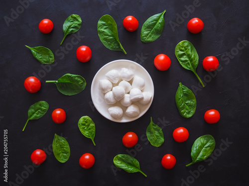 Italian mozzarella cheese, cherry tomatoes and spinach leaves on a black table. Pattern.
