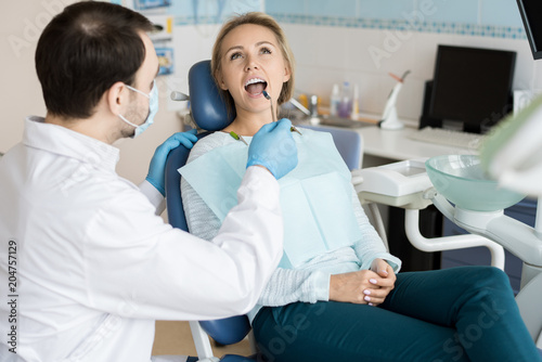 Doctor in gloves and mask exploring teeth of young girl with small mirror while sitting in chair. 