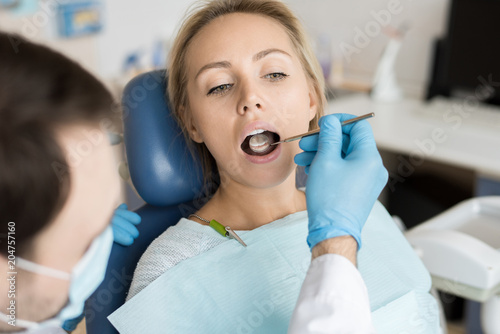 Anonymous dentist in gloves holding mirror tool and investigating teeth of young girl in chair. 