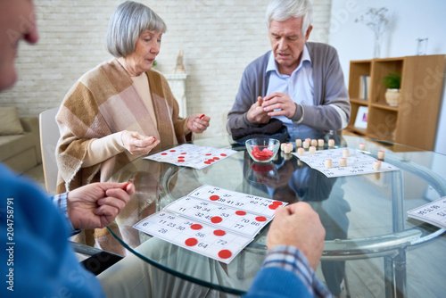 Fototapeta Naklejka Na Ścianę i Meble -  Senior patients of assisted living home gathering at table in living room and playing lotto at table. 