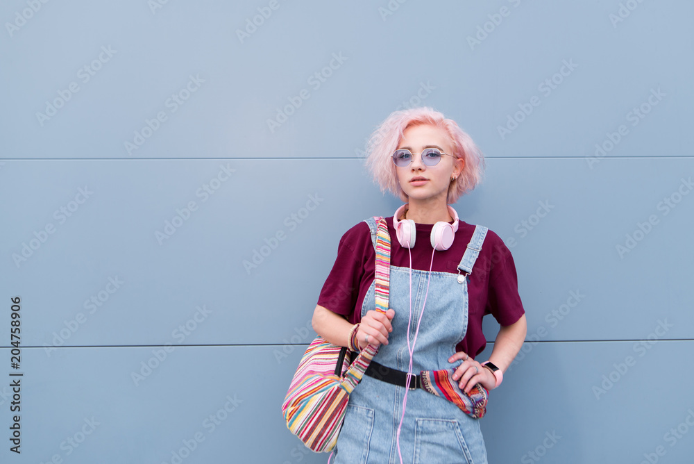 Portrait of a stylish bright young girl with headphones, sunglasses and colored hair on a blue background. Hipster girl on the background of a blue wall. Portrait of a stylish bright young girl with headphones, sunglasses and colored hair on a blue background. Hipster girl on the background of a blue wall.