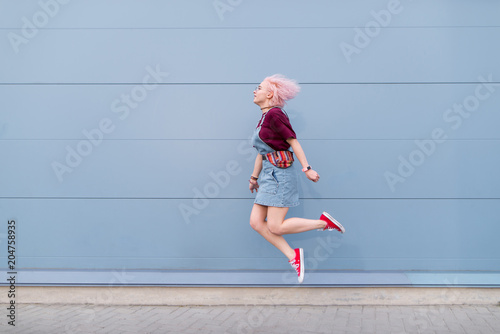 Fototapeta Naklejka Na Ścianę i Meble -  Positive girl in stylish clothes jumping against the background of a blue wall. A bright young woman jumping on a blue background.