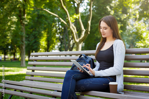 Motivated asian freelancer in the park. Girl on a bench, drinking coffee and working with tablet