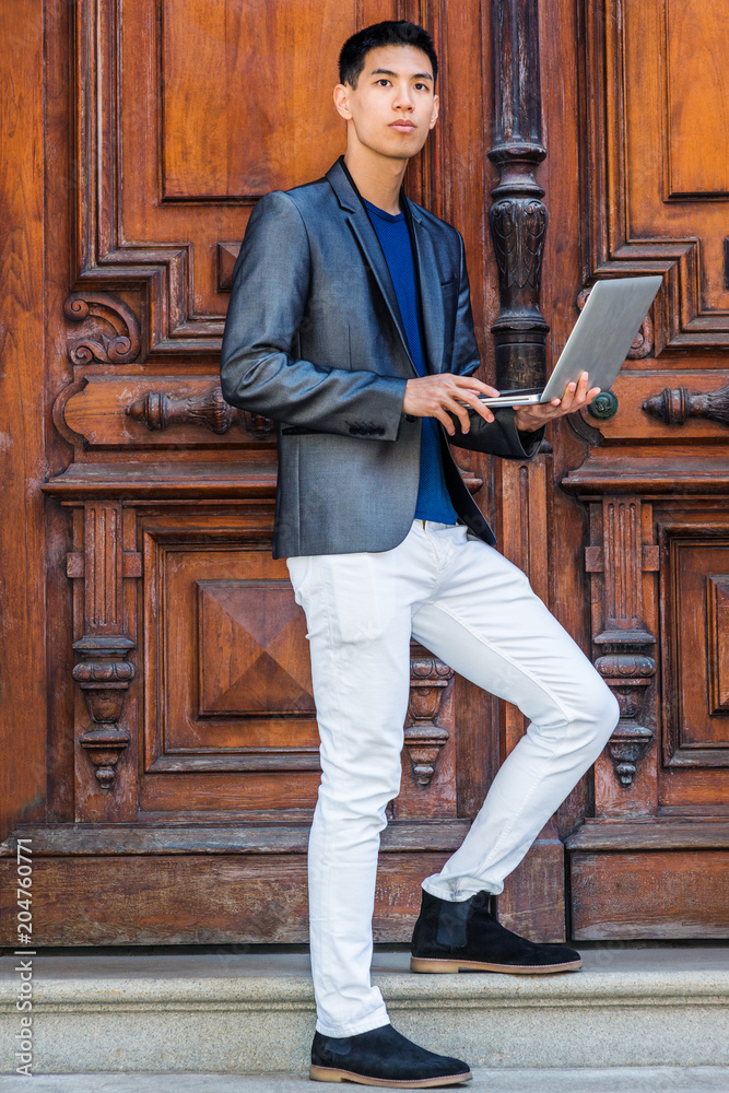 Young Asian American Man studying, working in New York, wearing fashionable gray blazer, white pants, black shoes, standing by old fashion style office door, working on laptop computer, looking away.