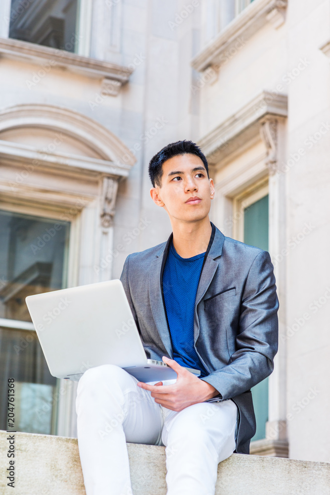 Young Asian American Man studying, working in New York, wearing gray blazer, white pants, sitting outside old office building on campus, working on laptop computer, looking up, thinking.