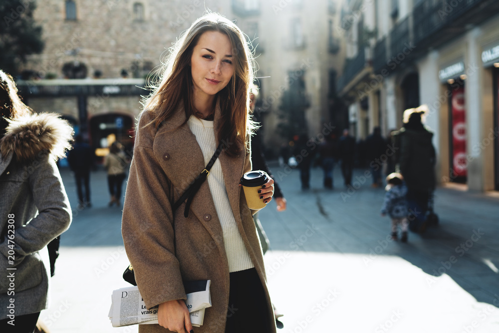 Freelancer female going to the coworking space with a coffee paper cup and a newspaper in her hand. Business woman wearing casual outfit relaxing outdoors with a cup of coffee during free time.