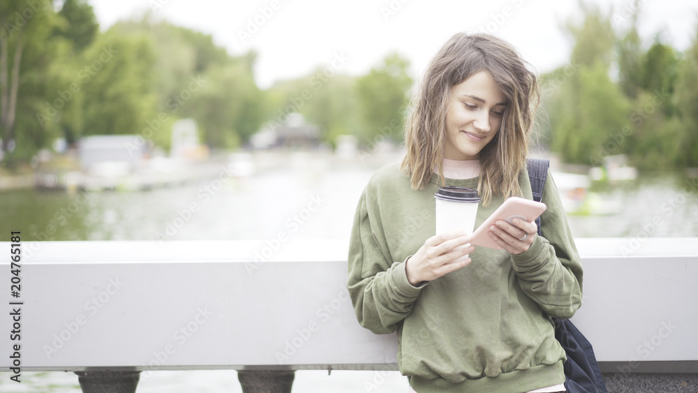 Fototapeta premium A smiling happy laughing young girl is listening to a music outdoors dressed in a green sweatshirt drinking a cup of coffee