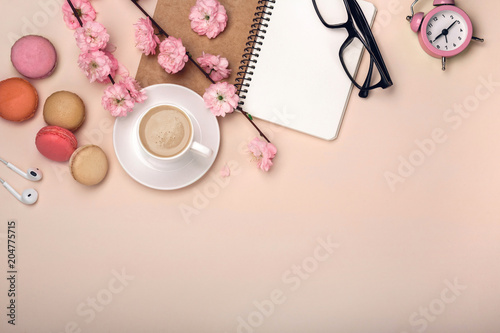 White cup with cappuccino, sakura flowers, macarons, alarm clock, notebook on a pastel pink background.