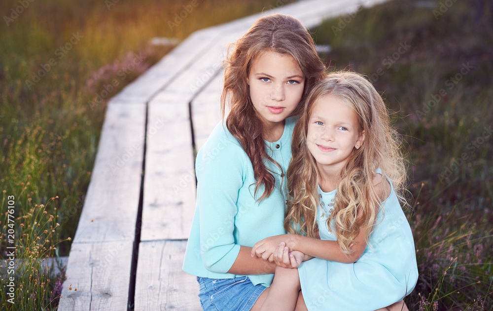 Fototapeta premium Sisters relaxing outdoors while sitting on a wooden path in a field at sunset.