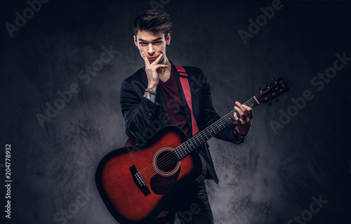 Handsome young thoughtful musician with stylish hair in elegant clothes posing with a guitar in his hands.