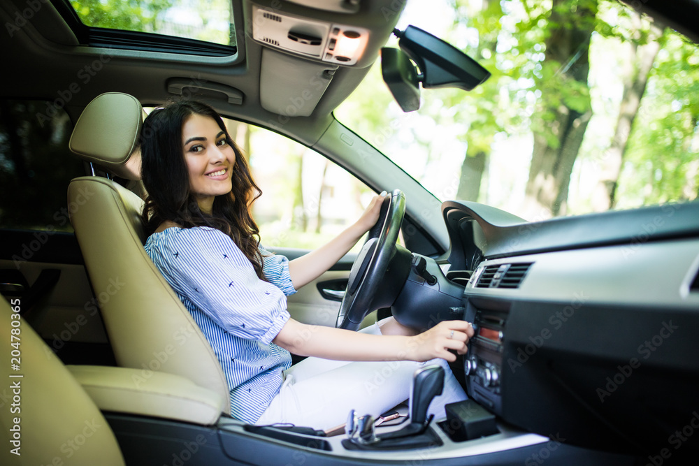 Young smiling woman driver with fastened safety seat belt turning on or tuning vehicle audio, finding a playlist for a pleasant and comfortable ride