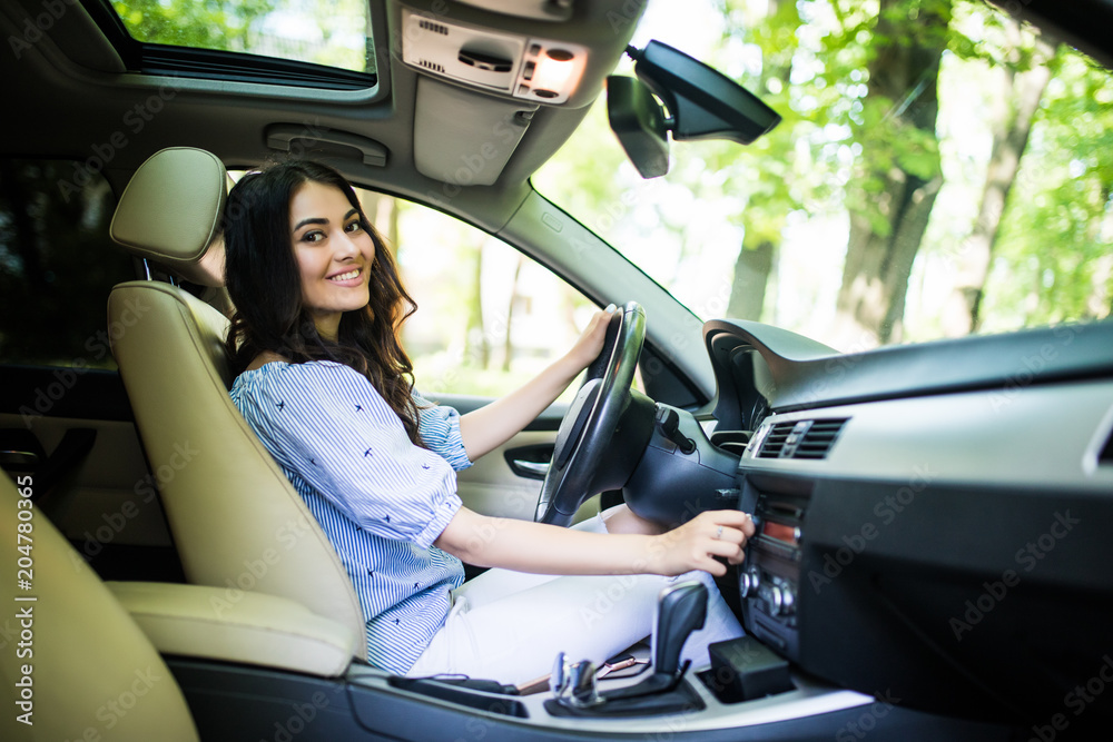 Naklejka premium Young smiling woman driver with fastened safety seat belt turning on or tuning vehicle audio, finding a playlist for a pleasant and comfortable ride