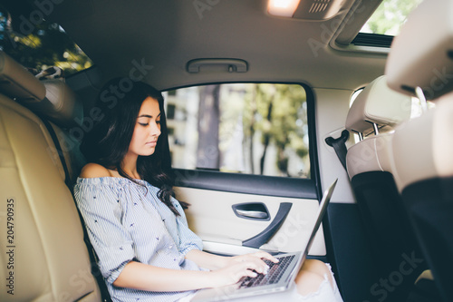 Beautiful business woman is using a laptop and smiling while sitting on back seat in the car