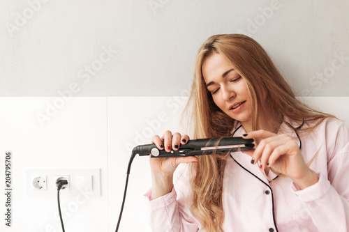 Close up photo of young lady sitting on bed and using hair straightener at home isolated