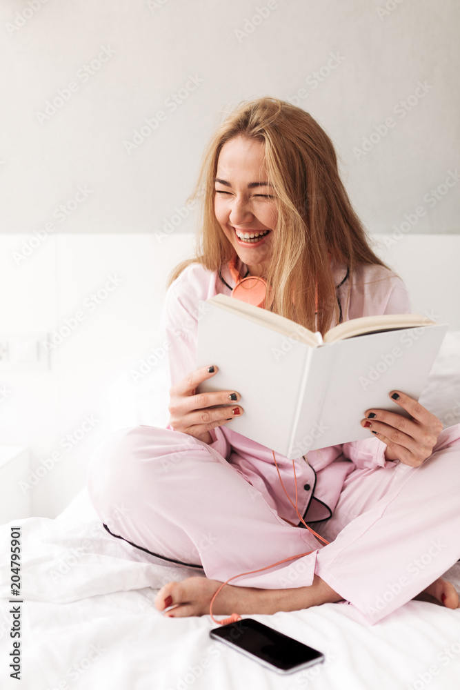 Young smiling lady sitting on bed with book in hands and happily laughing at home isolated Young smiling lady sitting on bed with book in hands and happily laughing at home isolated