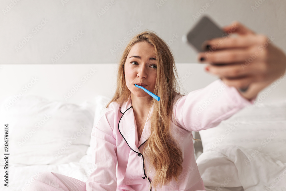 Portrait of young pretty lady sitting on bed with toothbrush and taking photos on cellphone at home isolated Portrait of young pretty lady sitting on bed with toothbrush and taking photos on cellphone at home isolated