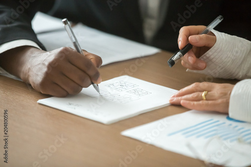 Diverse male and female partners playing tic tac toe or noughts and crosses game writing in notebook at office desk, marketing strategic tactical planning, business competition concept, close up view