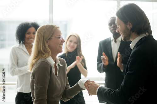 Boss handshaking rewarding motivating successful female employee congratulating with promotion  appreciating good work results  making partnership deal  expressing respect while colleagues applauding