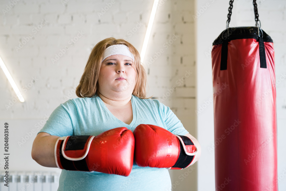 Waist up portrait of confident obese woman wearing boxing gloves posing looking at camera  ready for extreme training in gym, copy space