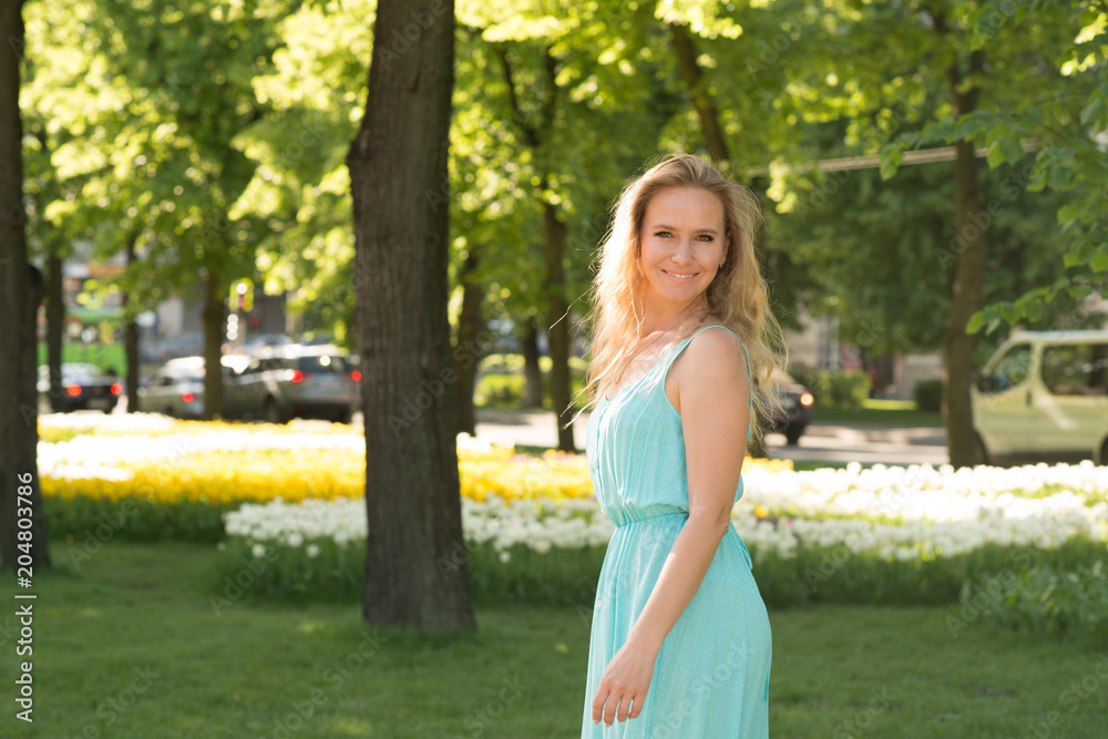 Waist up emotional portrait of the attractive blonde woman in a blue dress on the street against the background of flowers Waist up emotional portrait of the attractive blonde woman in a blue dress on the street against the background of flowers