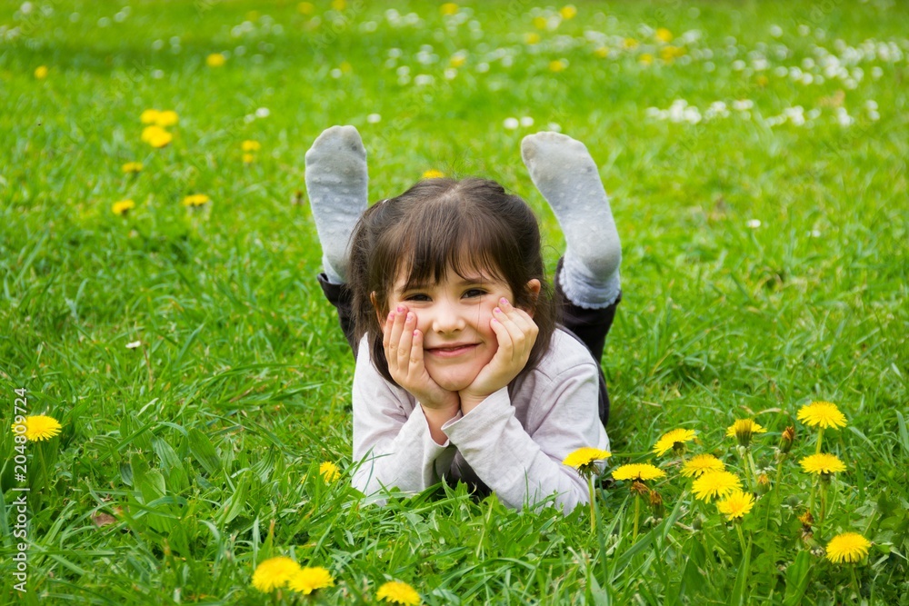 Happy little girl lying down on the grass with hands holding her face and feet up at park full of yellow flowers. Cute kid smiling on spring season Happy little girl lying down on the grass with hands holding her face and feet up at park full of yellow flowers. Cute kid smiling on spring season