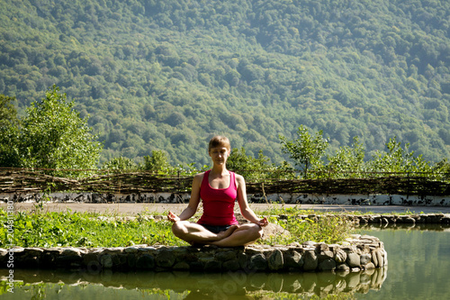 girl in shorts and t-shirt meditating on the shore of a mountain lake sitting by the water