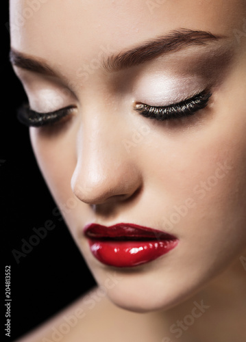 Portrait of young beautiful woman with evening make up touching her face, close eyes, over black background. Red lips and nails. Luxury skincare and fashion makeup concept, jewelery earnings.