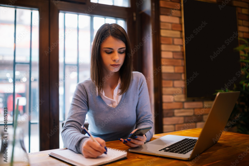 Young attractive brunette is writing down notes that she just received on her mobile phone Young attractive brunette is writing down notes that she just received on her mobile phone