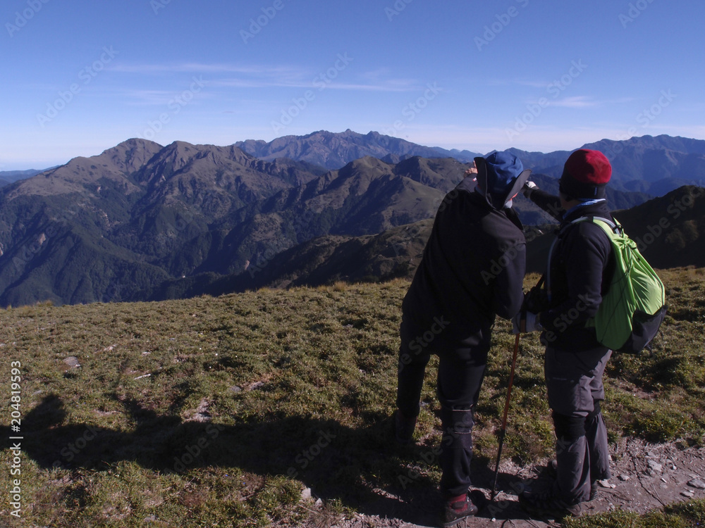 Two male hikers are pointing at the mountains far away and talking about what the names of those mountain are.