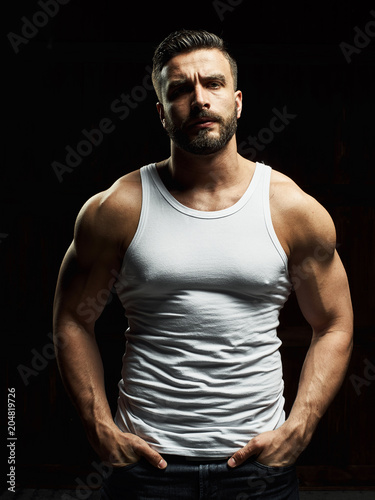 A strong, a serious, muscular man in a white t-shirt is worth and looks in camera. He's got the beard on the face and different emotions © Pavel