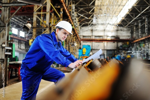Working engineer in a white construction helmet with a project or drawing plan on the background of an industrial plant