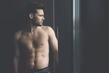 Feeling hopeless. Profile of young bearded shirtless man with developed muscularity is standing and looking through the window wistfully. Copy space in right side