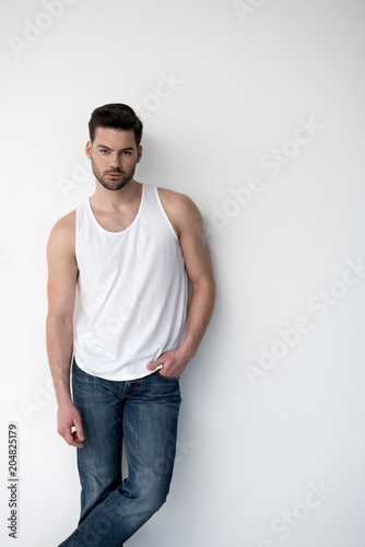 I am number one. Portrait of stylish pleasant guy wearing casual clothes is leaning on white wall and looking at camera confidently. He is holding hand in pocket of his jeans. Copy space