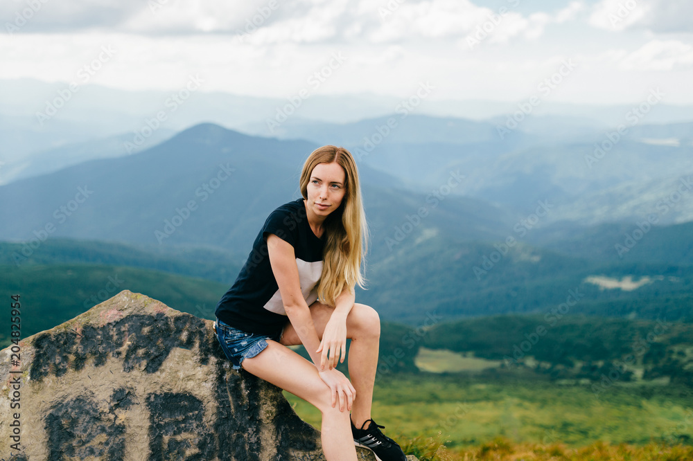 Young girl sitting on stone in mountains.  Mood portrait of long haired adorable blonde babe outdoor on vacation. Female hiker relaxing at nature. Beautiful landscape view. Cute lovely model posing.