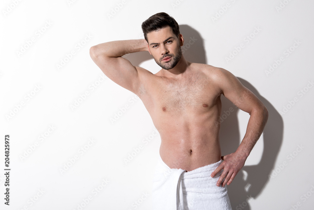 Portrait of pensive young bearded man is standing with towel on his hips and looking at camera thoughtfully. He is holding hand behind his head and other one on his hip. Copy space in the left side