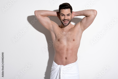 Enjoying my life. Portrait of masculine young smiling man with stubble is standing in towel and looking at camera with joy while holding hands behind head. Beauty concept