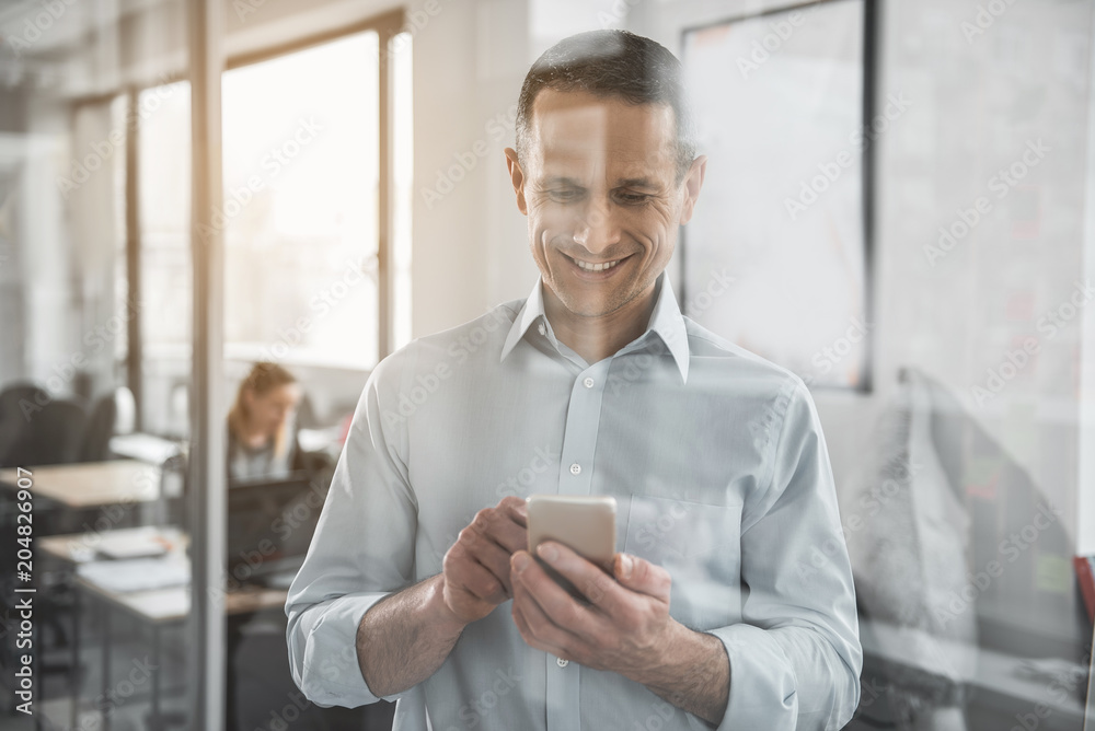 Portrait of cheerful businessman typing in phone while locating in office. Glad employer using appliance concept Portrait of cheerful businessman typing in phone while locating in office. Glad employer using appliance concept
