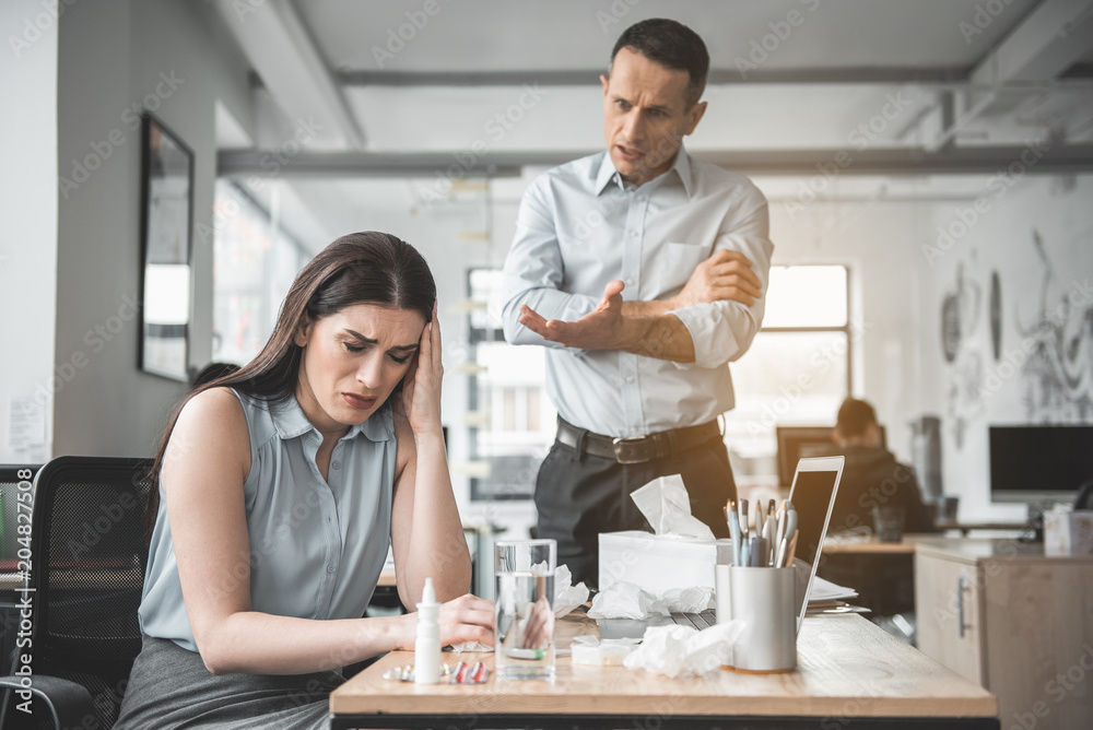 Portrait of unhappy ill lady sitting at table during job. Angry man arguing on her Portrait of unhappy ill lady sitting at table during job. Angry man arguing on her