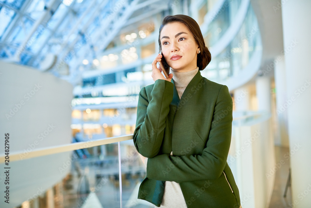 Waist up portrait of elegant Asian business lady speaking by phone standing on glass balcony in modern office building, copy space Waist up portrait of elegant Asian business lady speaking by phone standing on glass balcony in modern office building, copy space